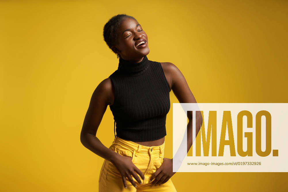 Positive young African American female in casual clothes with braided hair standing near yellow wall with hands in pockets JorgeMierteran_ID10736_325118_053