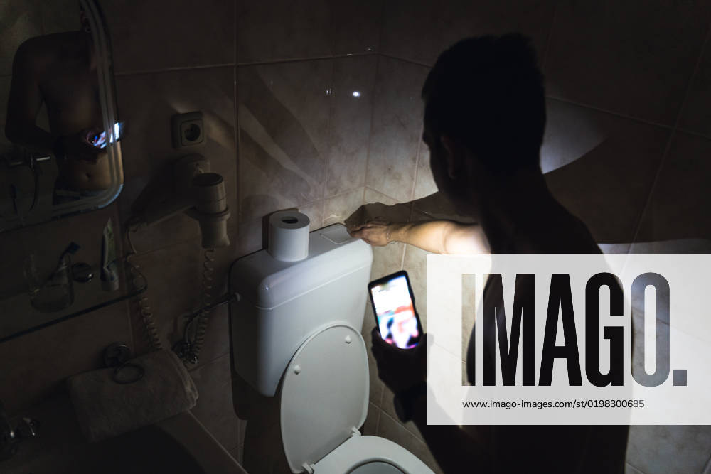 Stromausfall oder Blackout in der Wohnung. Ein junger Mann geht in einem dunklen Badezimmer auf die Toilette und leuchtet mit der Taschenlampe in seinem Smartphone. Energie Krise Symbolbild