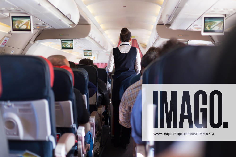 Interior of commercial airplane with passengers on seats during flight. Interior of commercial airplane with passengers on seats during flight. Stewardess in dark blue uniform walking the aisle. Horizontal composition.