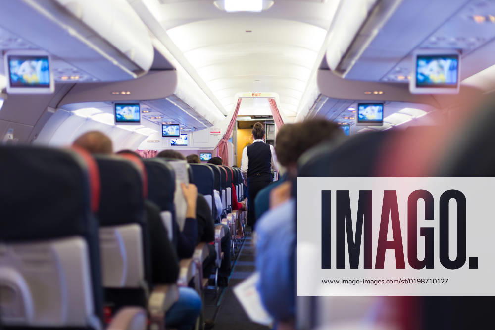Interior of commercial airplane with passengers on seats during flight. Interior of commercial airplane with passengers on seats during flight. Stewardess in dark blue uniform walking the aisle. Horizontal composition.