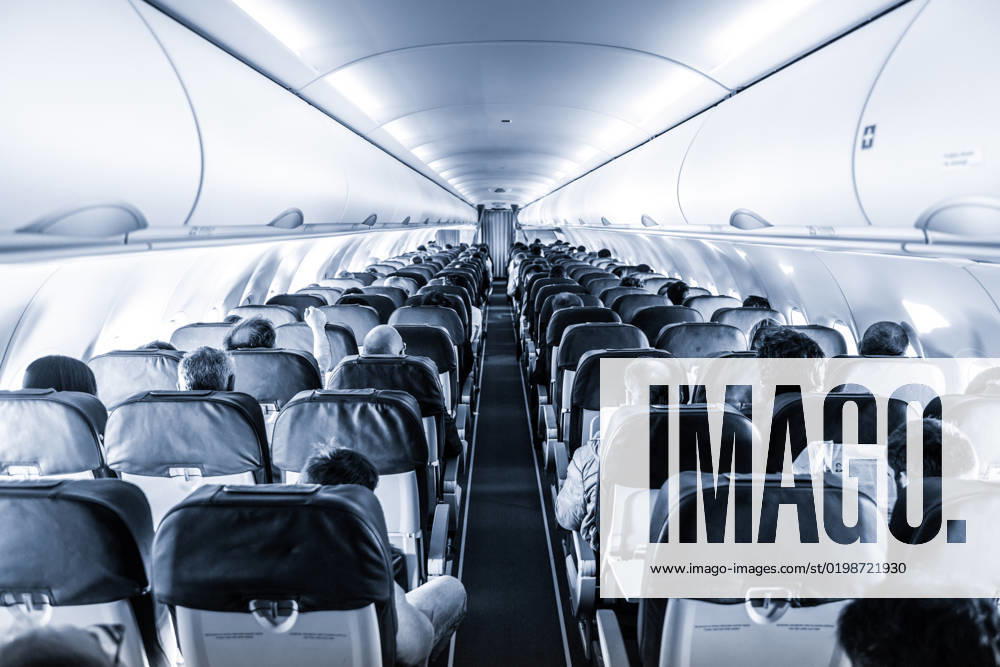 Interior of commercial airplane with passengers on their seats during flight. Interior of commercial airplane with unrecognizable passengers on their seats during flight shot from the rear of airplane. Black and white, blue toned image.