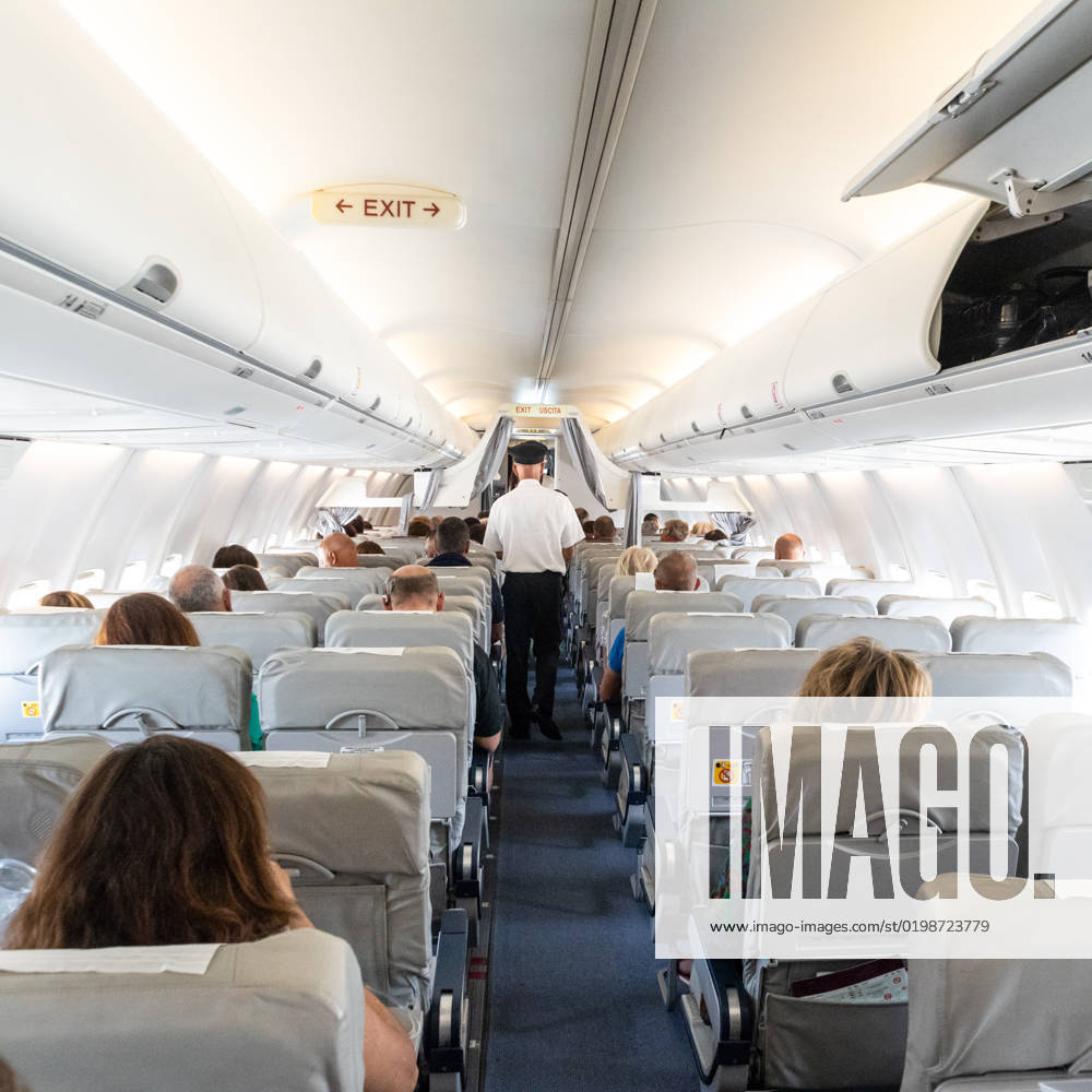 Interior of commercial airplane with passengers on their seats during flight. Interior of commercial airplane with unrecognizable passengers on their seats during flight. Steward in blue white uniform walking the aisle of commercial airplane.
