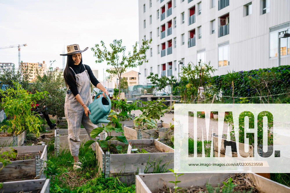 IMAGO Creative, Urban Lifestyle Smiling young woman wearing hat watering plants in urban garden model released, Symbolfoto, GDBF00022