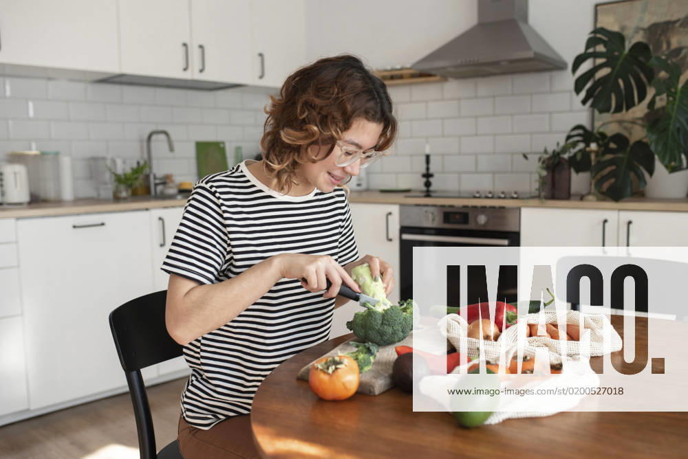 Smiling young woman cutting broccoli sitting at table in kitchen model released, Symbolfoto property released, VRAF00055