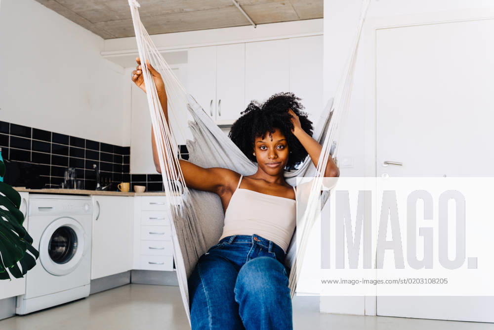 Positive young African American female in casual clothes with Afro hairstyle looking at camera while resting on hanging hammock near kitchen zone JakeJakab_ID190_6414_058