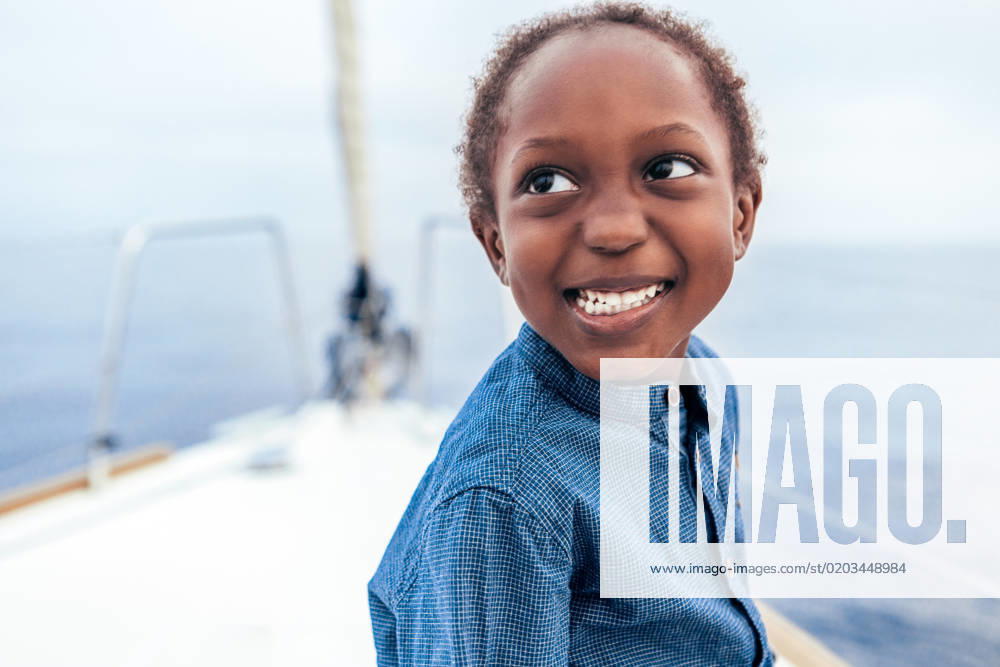Positive African American boy in casual shirt smiling and looking away while sitting on yacht against sea, Model released AlfonsoSaenz_ID12778_387457_041