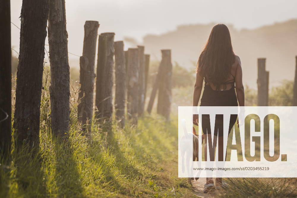 IMAGO Creative, Jahreszeit Sommer, Sommerferien Back view of unrecognizable female tourist with long dark hair in black dress walking on narrow path with dog during summer holidays in Asturias at sunset, Model released PabloMenendez_ID12520_379984_055