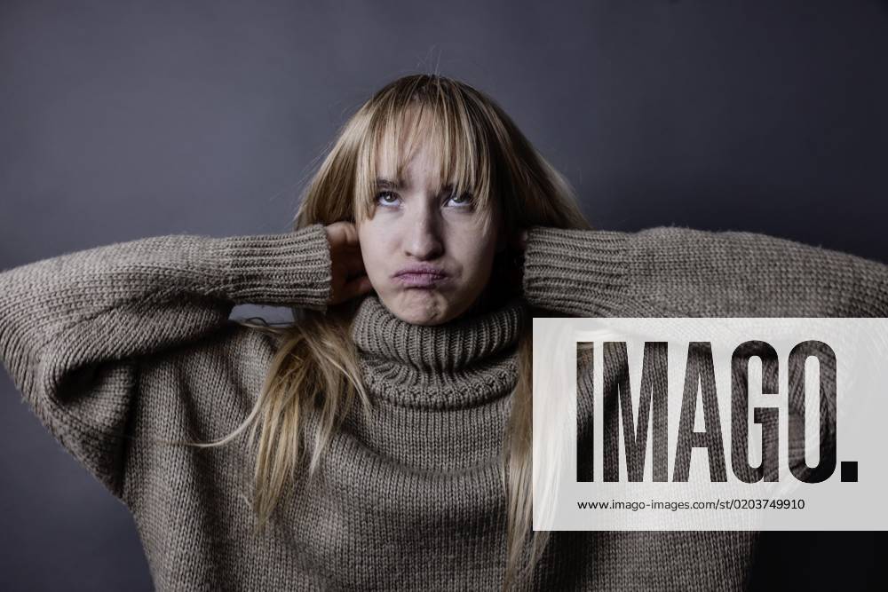 Junge Frau mit langem, blonden Haar und eimem Wollpullover verschränkt Arme hinter dem Kopf und verzieht das Gesicht, Portrait, Studioaufnahme
