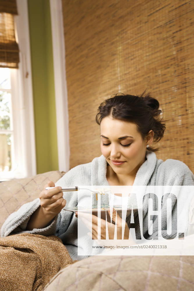 Young woman sitting on a couch in a bathrobe eating soup. Vertical shot.