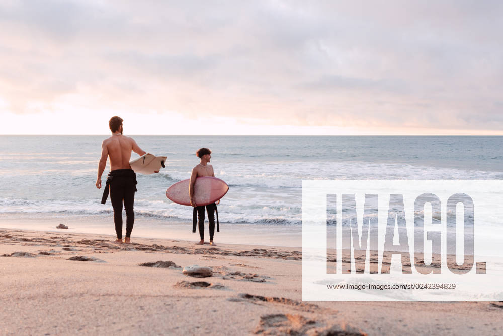 Distant male friends with surfboards and naked torso walking along wet sandy coast near waving sea and looking away during sunset, Model released NicoMortarini_ID13416_408702_012