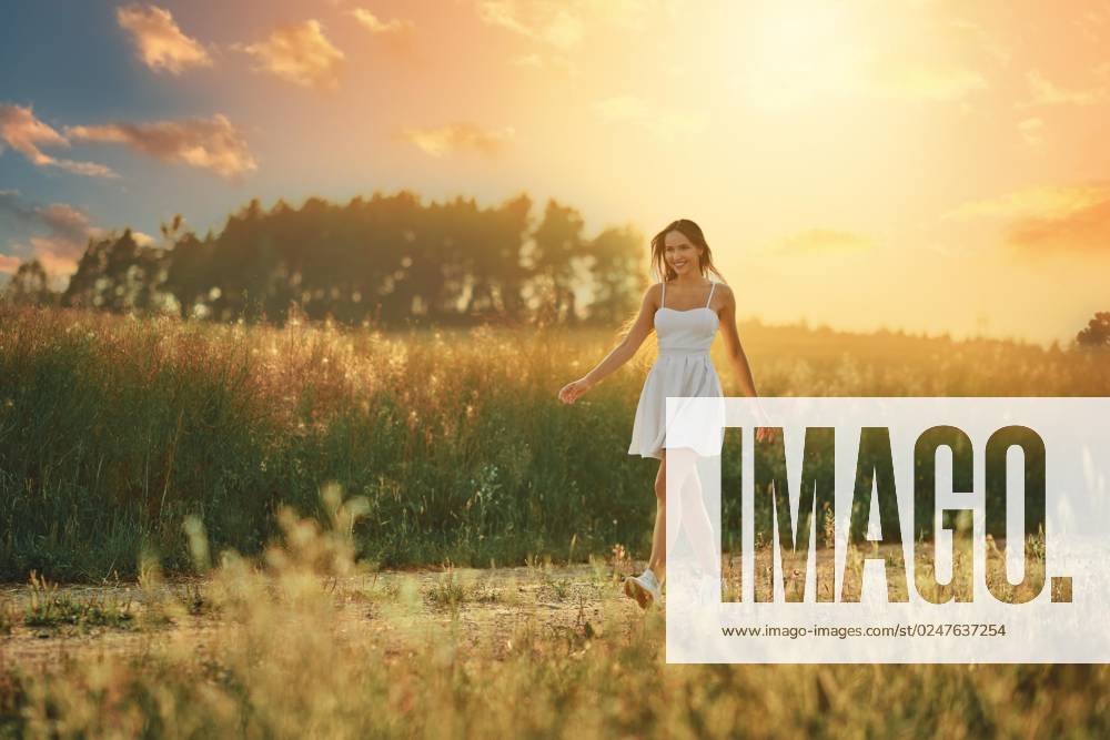 IMAGO Creative: Sommer, Abendspaziergang Glückliche junge Frau, die bei Sonnenuntergang auf einer Wiese entlang der Landstraße spaziert