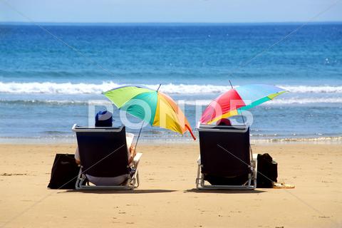 A couple relaxing on a beach under colorful umbrellas couple, people, romantic, person, love, two, pair, chair, chairs, beach, beaches, sand, sandy, water, sea, ocean, seashore, shore, coast, coastline, outside, outdoor, outdoors, summer, vacation, vacations, holiday, holidays, weekend, sit, sitting, relax, relaxing, umbrella, umbrellas, colorful, color, read, reading, view, back, happy, tropics, tropical, island, maine, yellow, blue, getaway, folding