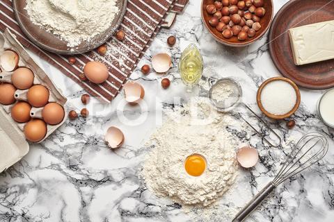 Close-up shot. Top view of a kitchenware and baking ingredients on a marble table background. There are some eggs and an eggshell, flour on a plate standing on a brown striped apron, sieve, butter, milk, sugar, whisk, oil in a sauce boat, scattered hazelnuts, flour on a table with yolk on it. flour, table, background, cooking, food, kitchen, pastry, preparation, white, egg, butter, top, above, bake, homemade, culinary, rural, home, basic, life, view, recipe, cookies, cuisine, ingredient, milk, wooden, sugar, decoration, yolk, hazelnuts, sweet, dessert, menu, pie, preparing, utensil, close-up, nobody, domestic, striped, apron, whisk, oil, plate, rustic, delicious, tasty, marble, kitchenware