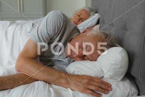 Front view of active senior Caucasian couple sleeping together in bed in bedroom, Staying at home in quarantine lockdown. Social distancing and self isolation stay at home,social distancing,quarantine,self isolation,real people,isolation,pandemic,distance,lock down,coronavirus,covid19,covid,Two People,Mature Adult,Woman,Female,Caucasian,Senior,Elderly,Man,Male,Mature Women,Caucasian Ethnicity,Senior Men,Senior Couple,Relationship,Married,Senior Citizen,Aged,Ageing Process,Retired,Retirement,Pensioner,Active Senior,Active,Casual Clothing,Grey Hair,Eyes Closed,Sleeping,Asleep,Relaxing,Rest,Resting,Bedtime,Home,House,Household,Homey,Apartment,Domicile,Abode,Authentic,Real Life,Comfortable,Bedroom,Bed,Pillow,Blanket,Together,Bonding,Closeness,Affection,Love,Care,Lifestyle,Domestic Life,Leisure,Indoors,YNGHRTP5BRIEF