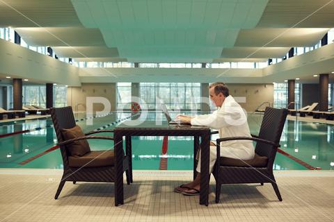 Full length side view of successful senior man using laptop while relaxing at luxury SPA resort with swimming pool in background, copy space {Table,Indoors,People,Men,Adult,Business,One Person,Sitting,Professional Occupation,Lifestyles,Healthcare And Medicine,Working,Expertise,Success,Businessman,Modern,Women,Business Person,Looking,Copy Space,Real People,Relaxation,Hospital,Restaurant,Serious,Hotel,Wood Material,Doctor,Thinking,Laptop,senior,mature,elderly,old,man,woman,successful,wealthy,rich,active,lifestyle,spa,resort,relax,swimming pool}