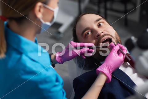 Smiling young man sitting in dentist chair while doctor examining his teeth {medical,woman,man,doctor,dentist,tool,specialist,procedure,patient,oral,gloves,examination,health,treatment,clinic,tooth,teeth,dental,medicine,caucasian,equipment,stomatology,visit,mouth,cabinet,checkup,cure,examine,young,uniform,surgical,protective,mask,toothache,latex,operation,hygiene,orthodontist,occupation,dentistry,workplace,indoors,instrument,sterile,heal,hospital,inspection}