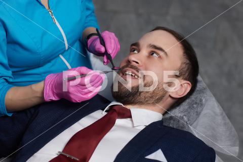 Smiling young man sitting in dentist chair while doctor examining his teeth {medical,woman,man,doctor,dentist,tool,specialist,procedure,patient,oral,gloves,examination,health,treatment,clinic,tooth,teeth,dental,medicine,caucasian,equipment,stomatology,visit,mouth,cabinet,checkup,cure,examine,young,uniform,latex,indoors,orthodontist,sterile,protective,mask,dentistry,hospital,toothache,surgical,instrument,hygiene,workplace,heal,operation,inspection,occupation}