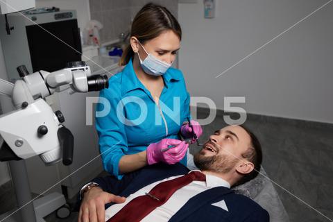 Smiling young man sitting in dentist chair while doctor examining his teeth {medical,woman,man,doctor,dentist,tool,specialist,procedure,patient,oral,gloves,examination,health,treatment,clinic,tooth,teeth,dental,medicine,caucasian,equipment,stomatology,visit,mouth,cabinet,checkup,cure,examine,young,uniform,surgical,protective,mask,toothache,latex,operation,hygiene,orthodontist,occupation,dentistry,workplace,indoors,instrument,sterile,heal,hospital,inspection}