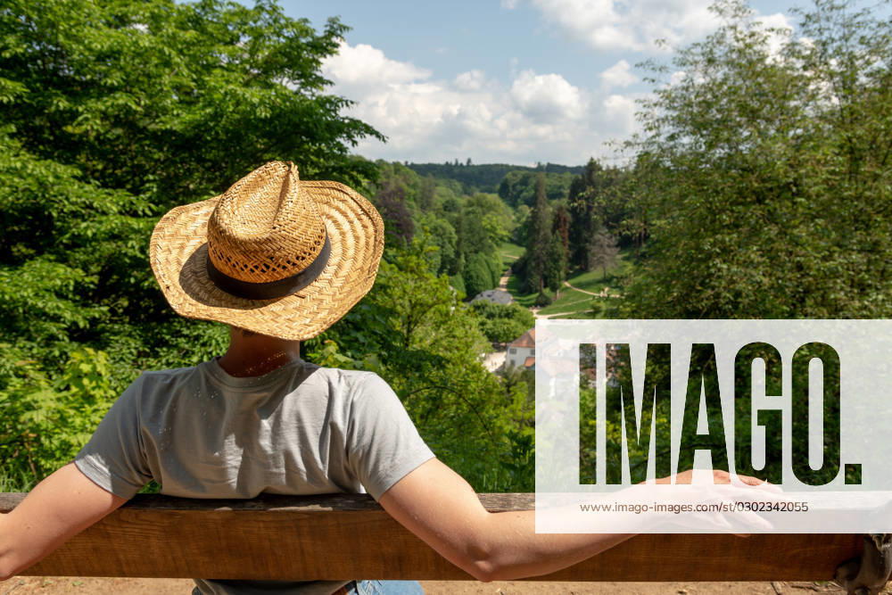 Woman with straw hat sitting on a park bench in the sun, looking at Staatspark Fürstenlager, Auerbach, Bensheim Woman with straw hat, gray t-shirt and jeans sitting on a wooden park bench in the sun, looking at Staatspark Fürstenlager, Auerbach, Bensheim, Germany ,model released, Symbolfoto