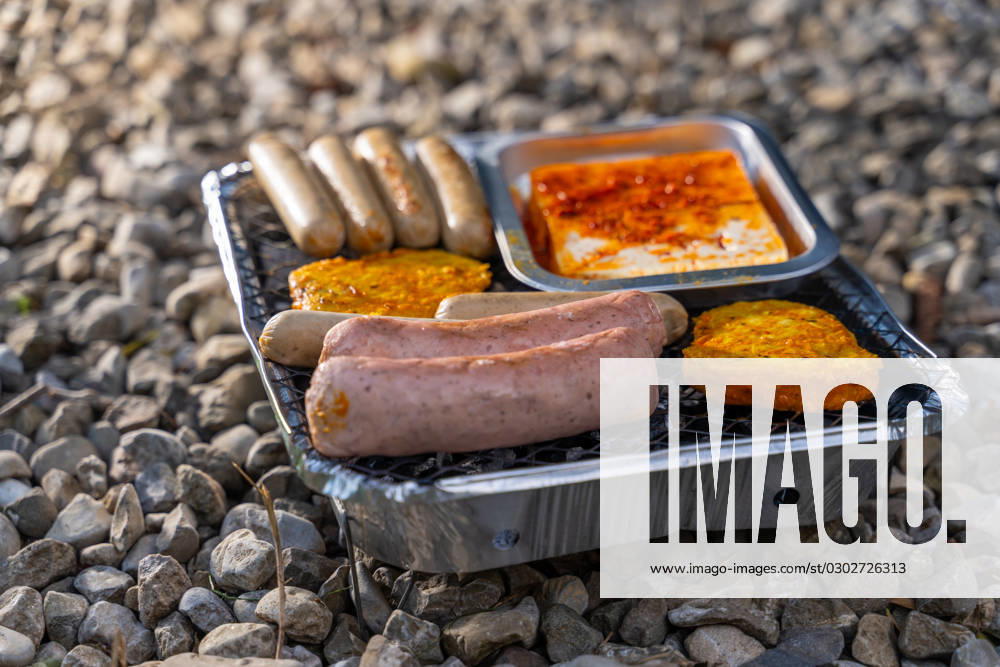 Vegetarisch Grillen, Bratwürste Vegan und Vegetarisch auf einem Einweggrill auf Kieselsteinen neben einer Wiese. Fleisch und Wurst Ersatzprodukte