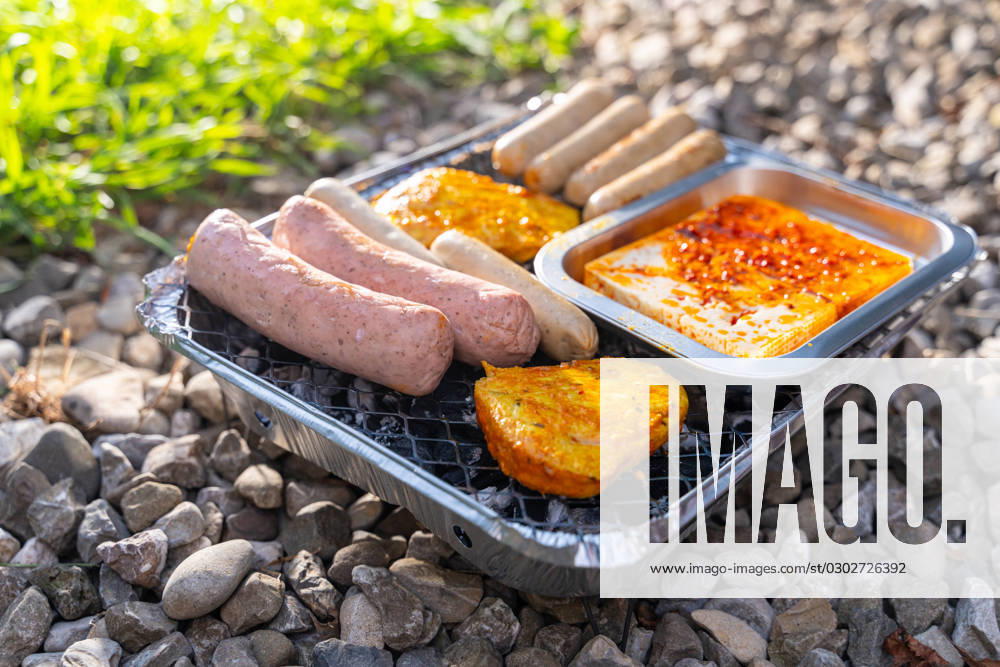 Vegetarisch Grillen, Bratwürste Vegan und Vegetarisch auf einem Einweggrill auf Kieselsteinen neben einer Wiese. Fleisch und Wurst Ersatzprodukte