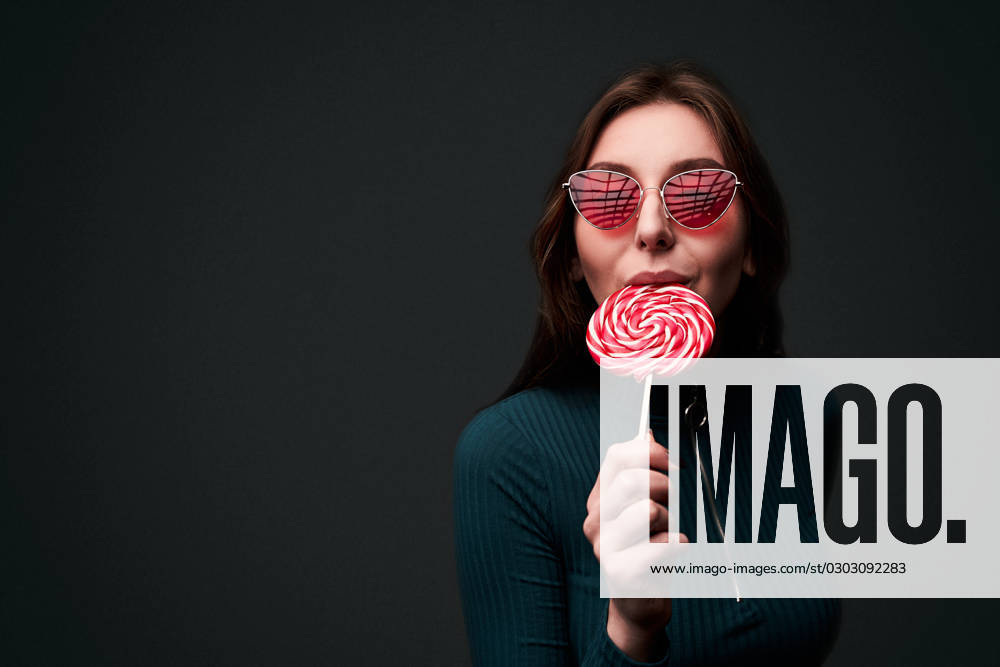 Portrait of a young beautiful stylish girl wearing red drop shaped sunglasses holding a red lollipop and biting it in a seductive sexy way isolated on dark grey background Portrait of a young beautiful stylish girl wearing red drop shaped sunglasses holding a red lollipop and biting it in a seductive sexy way ,model released, Symbolfoto