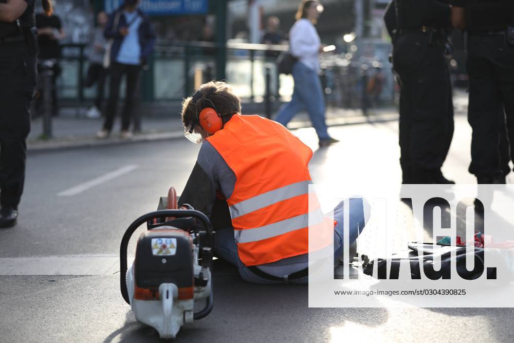 Berlin, Deutschland, 18.09.2023: Protest von Klimaaktivisten der Letzten Generation am Kottbusser Tor in Berlin-Kreuzberg. Einem Aktivisten wurden Ohrschutz, Augenschutz und Atemschutzmaske aufgesetzt, er wartet darauf, dass er mit einer Flex vom Straßenbelag abgelöst wird.