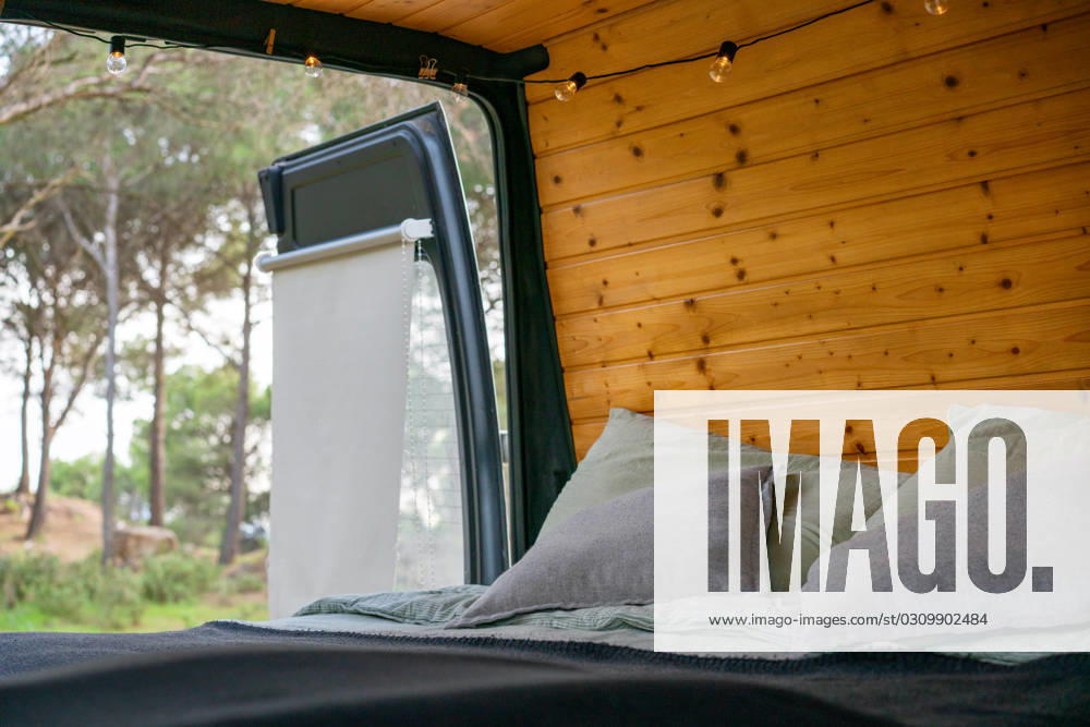 Tourist van with cozy bed pillows blanket near wooden planks wall ornament lights opened door with sun shade against blurred Andalusia trees Model Released LuisPina_ID16420_499228_018