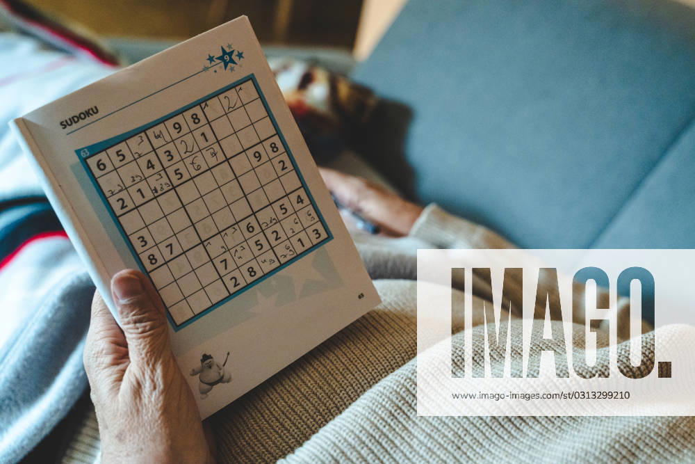 An old woman lies on a sofa in the living room and plays Sudoku in a book with a pen