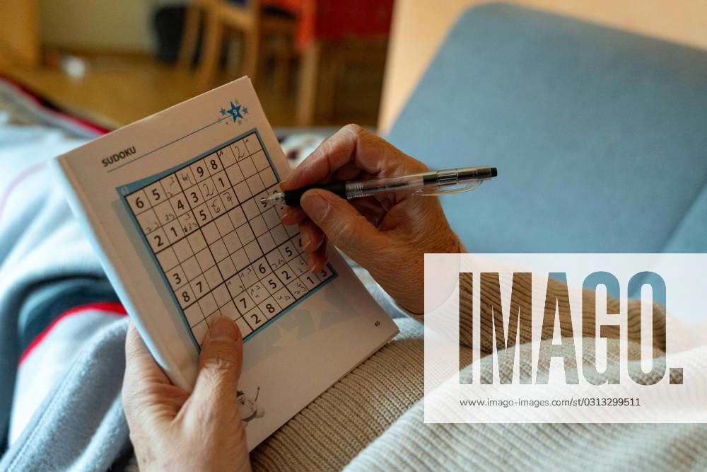 An old woman lies on a sofa in the living room and plays Sudoku in a book with a pen