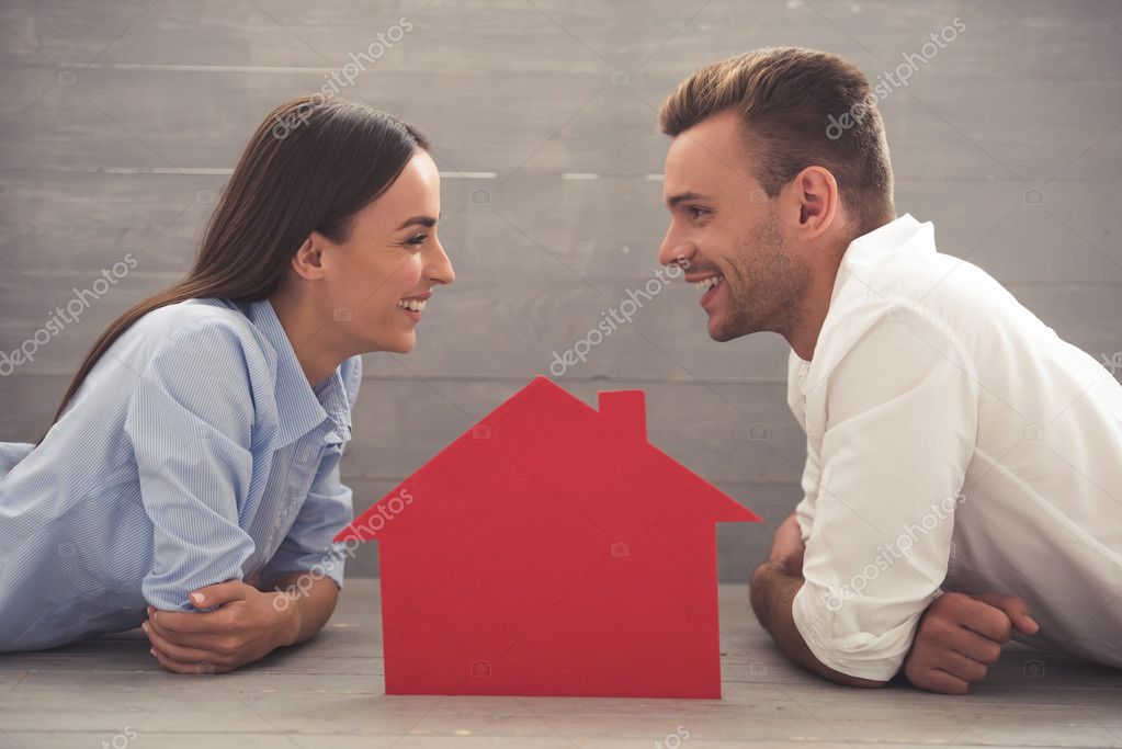Side view of beautiful young couple in classic shirts looking at each other and smiling while lying on the floor at home. A paper house between them model released