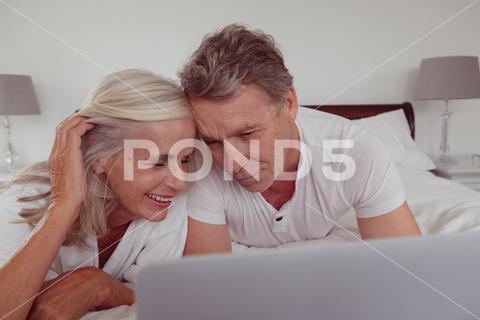 Front view of active senior Caucasian couple using laptop while lying on bed in bedroom at comfortable home, Staying at home in quarantine lockdown. Social distancing and self isolation stay at home, social distancing, quarantine, self isolation, real people, isolation, pandemic, distance, lock down, coronavirus, covid19, covid, Two People, Senior, Elderly, Woman, Female, Caucasian, Man, Male, Senior Women, Caucasian Ethnicity, Senior Men, Senior Couple, Relationship, Married, Senior Citizen, Aged, Ageing Process, Retired, Retirement, Pensioner, Active Senior, Active, Casual Clothing, Grey Hair, Lying, Relaxing, Talking, Discussing, Smiling, Happy, Cheerful, Joy, Using Laptop, Electronic, Wireless Technology, Communication, Connection, Home, House, Household, Homey, Apartment, Domicile, Abode, Authentic, Real Life, Comfortable, Bedroom, Bed, Pillow, Together, Bonding, Closeness, Affection, Love, Care, Lifestyle, Domestic Life, Leisure, Indoors, YNGHRTP5BRIEF