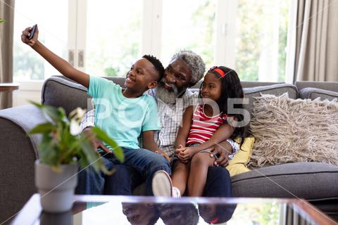 Senior African American man and his grandchildren enjoying their time at home together, sitting on a couch, embracing, smiling, using a smartphone, taking a selfie Two People, Child, Girl, Female, Mixed-Race, Mid Adult, Man, Male, Black, People, Family, Grandfather, Granddad, Granddaughter, Grandson, Siblings, Brother, Sister, Childhood, Together, Closeness, Interacting, Sitting, Couch, Holding, Using, Phone, Mobile Phone, Smartphone, Connection, Communication, Taking, Photo, Photograph, Selfie, Real People, Home, House, Domestic Life, Lifestyle, Enjoying, Free Time, Leisure, Relaxing, Weekend, Summer, Sunny, Love, Indoors