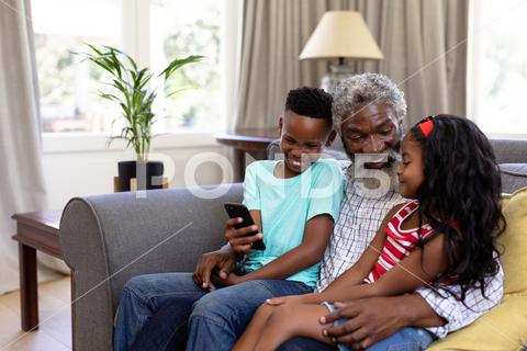 Senior African American man and his grandchildren enjoying their time at home together, sitting on a couch, embracing, smiling, using a smartphone Three People, Child, Girl, Female, Mixed-Race, Boy, Male, Black, Mid Adult, Man, People, Family, Grandfather, Granddad, Granddaughter, Grandson, Siblings, Brother, Sister, Childhood, Together, Closeness, Interacting, Sitting, Couch, Holding, Using, Phone, Mobile Phone, Smartphone, Connection, Communication, Real People, Home, House, Domestic Life, Lifestyle, Enjoying, Free Time, Leisure, Relaxing, Weekend, Summer, Sunny, Love, Indoors