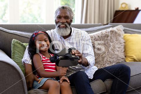 Senior African American man and his granddaughter enjoying their time at home together, sitting on a couch, holding a VR headset, looking at the camera and smiling Two People, Child, Girl, Female, Mixed-Race, Mid Adult, Man, Male, Black, People, Family, Grandfather, Granddad, Granddaughter, Childhood, Together, Closeness, Interacting, Sitting, Couch, Happy, Smiling, Holding, Vr, Virtual Reality, Headset, Connection, Communication, Real People, Home, House, Domestic Life, Lifestyle, Enjoying, Free Time, Leisure, Relaxing, Weekend, Summer, Sunny, Love, Indoors, Looking At Camera