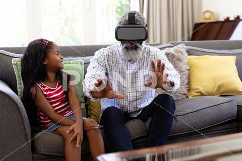 Senior African American man and his granddaughter enjoying their time at home together, sitting on a couch, wearing a VR headset Two People, Child, Girl, Female, Mixed-Race, Mid Adult, Man, Male, Black, People, Family, Grandfather, Granddad, Granddaughter, Childhood, Together, Closeness, Interacting, Sitting, Couch, Happy, Smiling, Wearing, Using, Vr, Virtual Reality, Headset, Connection, Communication, Real People, Home, House, Domestic Life, Lifestyle, Enjoying, Free Time, Leisure, Relaxing, Weekend, Summer, Sunny, Love, Indoors