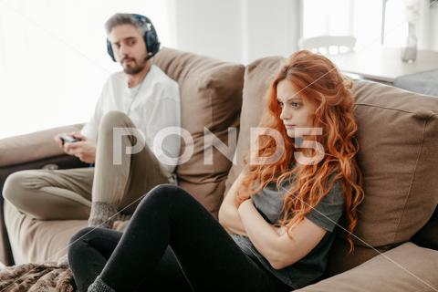Full body of sad redhead female sitting on comfortable couch near boyfriend playing interesting videogame in light living room at home couple, videogame, bored, ignore, pastime, leisure, relationship, play, hobby, redhead, sad, together, home, ginger, spend time, upset, boyfriend, girlfriend, long hair, apartment, light, man, woman, flat, red hair, at home, soulmate, entertain, boredom, partner, sofa, living room, couch, headphones, game, weekend, free time, gamepad, chill, problem, gamer, player, frustrate, settee, trouble, disappoint, inside
