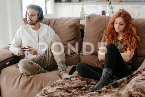 Full body of sad redhead female sitting on comfortable couch near boyfriend playing interesting videogame in light living room at home couple, videogame, bored, ignore, pastime, leisure, relationship, play, hobby, sad, together, home, spend time, upset, boyfriend, girlfriend, apartment, light, man, woman, flat, at home, soulmate, entertain, boredom, partner, sofa, living room, couch, headphones, game, weekend, free time, gamepad, chill, problem, gamer, player, frustrate, settee, trouble, disappoint, inside, full body, redhead, ginger, long hair, red hair