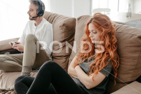 Full body of sad redhead female sitting on comfortable couch near boyfriend playing interesting videogame in light living room at home couple, videogame, bored, ignore, pastime, leisure, relationship, play, hobby, sad, together, home, spend time, upset, boyfriend, girlfriend, apartment, light, man, woman, flat, at home, soulmate, entertain, boredom, partner, sofa, living room, couch, headphones, game, weekend, free time, gamepad, chill, problem, gamer, player, frustrate, settee, trouble, disappoint, inside, redhead, ginger, long hair, red hair