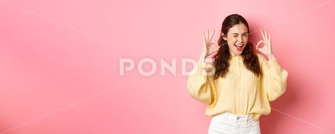 Portrait of cheerful pretty girl winking and saying yes, showing okay sign, alright gesture, praise good job, well done, standing pleased against pink background. student, girl, background, okay, good, approve, alright, agree, yes, praise, woman, advertising, fashion, beauty, person, portrait, grinning, joy, pink, beautiful, joyful, pleased, positive, smile, stylish, studio, horizontal, young, gesturing, glamour, holiday, excited, shopping, isolated, adult, girlfriend, advertisement, casual, smiling, emotive, slim, upbeat, glamorous, cheerful, emotions, excitement, fashionable, brunette