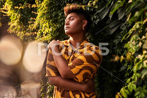 Dreamy young Filipino non binary man wearing cool shirt with earring embracing himself with love standing with eyes closed in green garden {man,self love,accept,imperfection,lgbt,non binary,garden,alternative,embrace,self,tolerance,identity,lgbtq,city,park,sunglasses,sunlight,generation,cool,daydream,queer,asian,filipino,personality,unaltered,pensive,complexion,eyes closed,contemporary,eyewear,ethnic,individuality,calm,guy,shirt,summer,independent,hairstyle,haircut,informal,outdoors,blur,accessory}