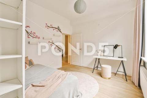 A bedroom with a bed, desk and book shelf in the room is white and has pink flowers on it {room,bedroom,house,furniture,home,flowers,bed,desk,floor,modern,lamp,table,sofa,comfortable,decor,living,apartment,luxury,window,wood,architecture,design,residential,indoors,light,hotel,pillow,carpet,chair,decoration,relaxation,wall,inside,domestic,residence,relax,corner,living room,rest}