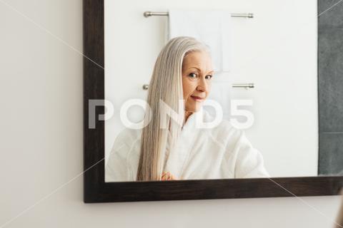 Portrait of an aged woman with long grey hair. Female looking in the mirror admires her hair. admires, adult, aged, bathrobe, care, caucasian, grey, grey hair, indoors, lifestyle, long hair, looking, mature, mirror, morning, one, people, person, portrait, real, reflection, routine, senior, woman