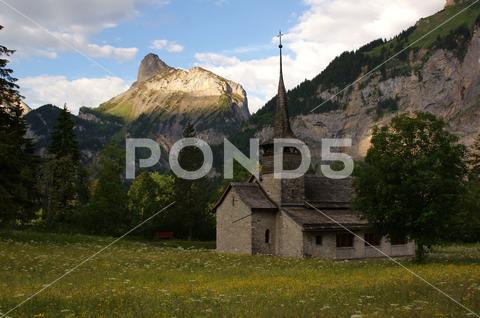 A scenic view of an old church located in a field surrounded by mountains in Kandersteg, Switzerland ancient, architecture, background, building, church, clouds, field, foliage, grass, green, leaves, meadow, mountains, old, religion, scenic, sky, trees