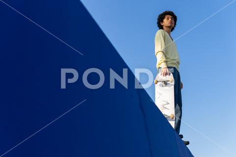 Low angle full body of thoughtful young Asian male skater in casual outfit standing on ramp with skateboard and looking away against blue cloudless sky man, skateboard, skater, ramp, thoughtful, hobby, skate park, activity, pensive, blue sky, male, young, casual, ethnic, subculture, urban, asian, serious, millennial, extreme, cool, confident, recreation, individuality, summer, street style, personality, sporty, daytime, cloudless, ponder, peaceful, carefree, calm, balance, full body, looking away, full length, low angle, from below