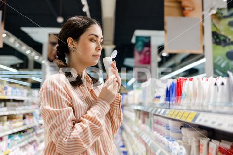 Portrait of Caucasian woman holds cream tube and sniffs fragrance. Choice of care cosmetics in supermarket. Concept of shopping and purchase. cosmetic, cream, tube, sniff, supermarket, cosmetics, care, purchase, product, shopping, woman, choice, close up, shampoo, perfume, shelf, smell, holding, buy, store, beautiful, concept, caucasian, consumer, customer, holds, skin, tester, person, retail, female, one, shopaholic, bottle, young, consumerism, ingredients, lifestyle, mall, portrait, casual, side view, hand, package, market, brunette, looking, shop, corridor, sale