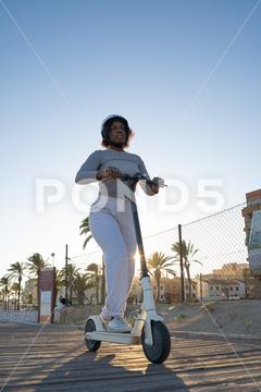 From below pensive young African American female in casual clothes and protective helmet looking away while riding electric scooter on city street with palm trees {woman,scooter,electric,city,helmet,ride,street,vehicle,pensive,transport,ethnic,urban,african american,female,palm trees,casual,young,black,safety,protect,modern,commute,town,contemporary,daytime,activity,thoughtful,curly hair,road,serious,summer,safe,confident,district,daylight,active,pavement,dreamy,brunette,blurred background,looking away,from below,low angle}