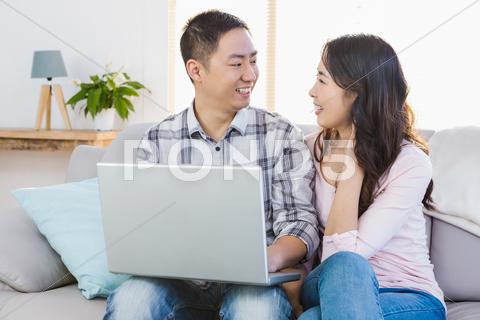 Happy young couple using laptop while sitting on sofa at home 30s, Mid Adult, Man, Male, Chinese, Young Adult, Woman, Female, Love, Feelings, Affection, Fondness, Lover, Relationship, Closeness, Romance, Romantic, Holding, Couple, Casual, Togetherness, Affectionate, Girlfriend, Heterosexual, Fashion, Cool, Using, Typing, Laptop, Notebook, Technology, Computer, Wireless, Modern, Wireless Technology, Connection, Communication, Happy, Smiling, Cheerful, Showing, Siting, Sofa, Relaxation, Home, Confidence, Face To Face, Indoors, REUNITEDP5BRIEF