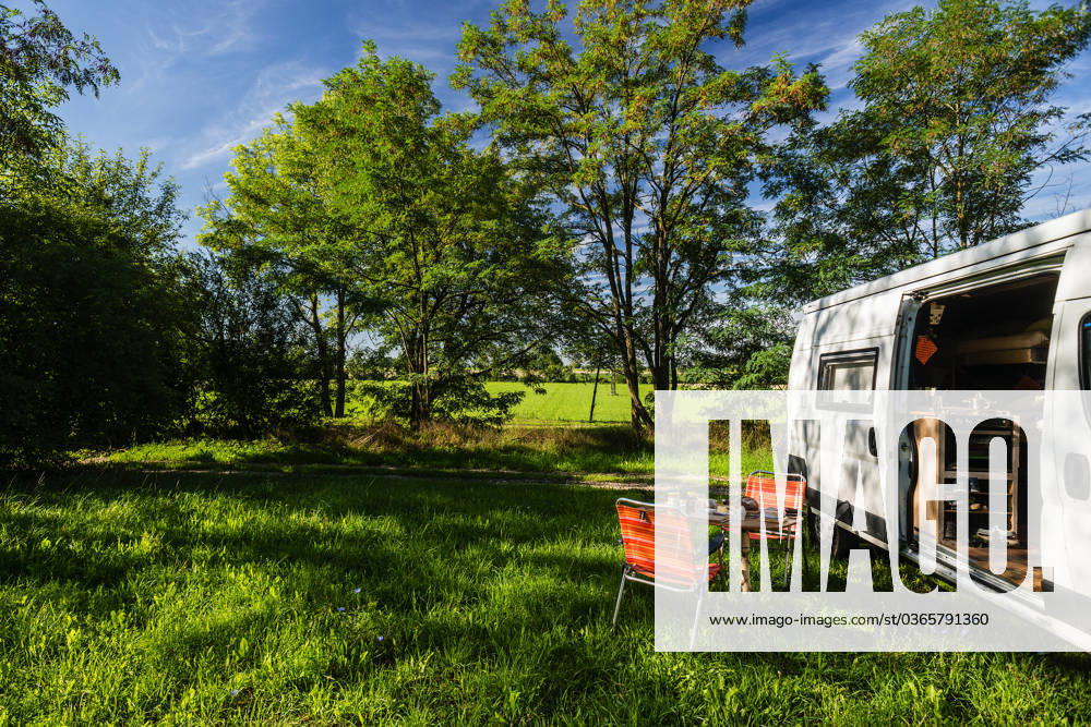 Ein Camper steht zwischen Bäumen an einem sonnigen Sommertag auf einer Wiese im Grünen, ein gedeckter Frühstückstisch mit Stühlen steht vor der Schiebetür des Campers. Regensburg, Bayern, Deutschland. In der Altstadt von Regensburg