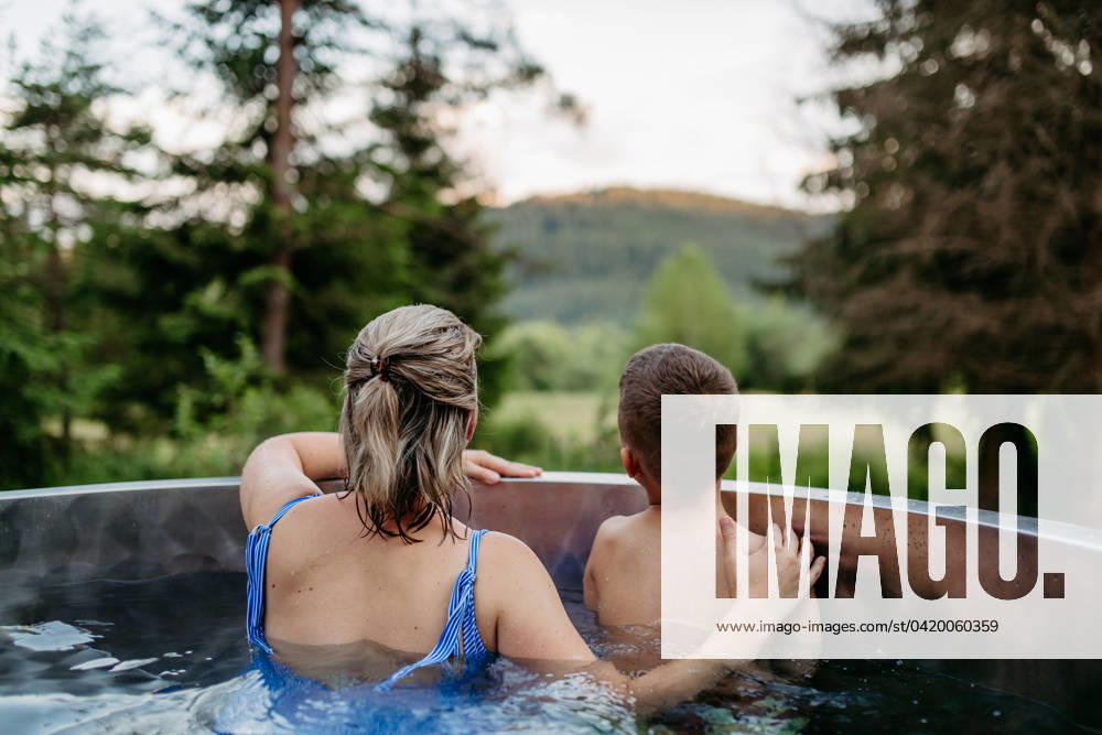 IMAGO Creative, Jahreszeit Sommer, Hitze Mother with her little son enjoying view at nature while bathing in wooden barrel hot tub in the terrace of the cottage. Mother with her little son enjoying view at nature while bathing in wooden barrel hot tub in the terrace of the cottage. Wooden bathtub with a fireplace to burn wood and heat water. model released